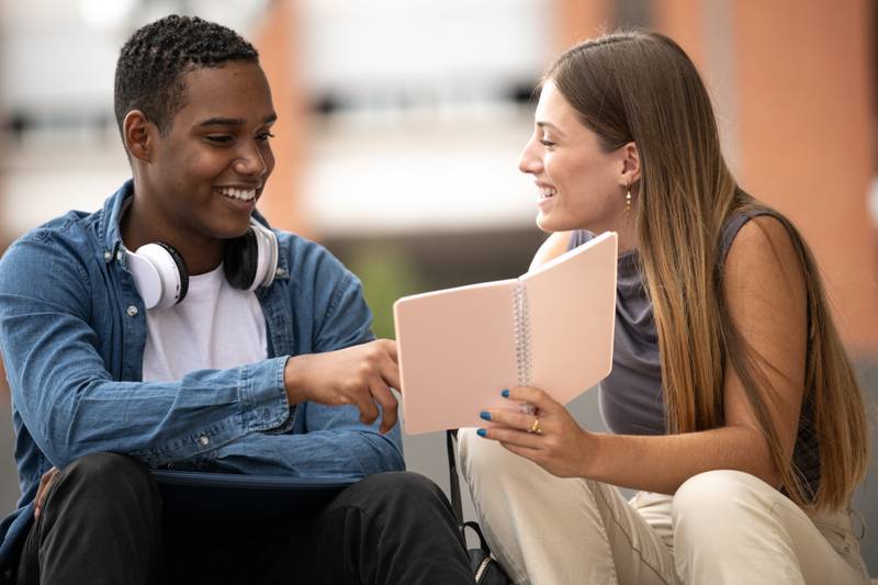 Young beautiful couple smiling and reading together a notebook.Two diverse students sitting outside talking while holding files.