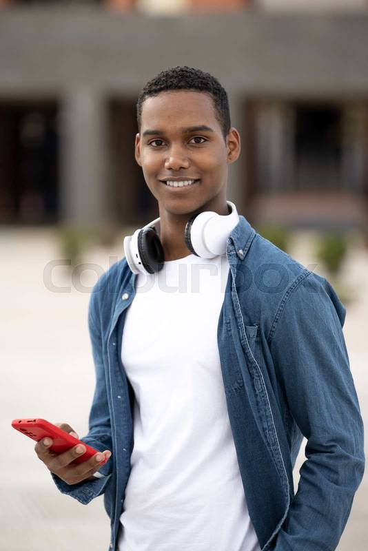 Young handsome guy smiling and looking at camera relaxed and using his phone.Satisfied man smiling and standing and texting while wearing headphones.