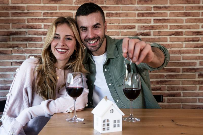 Young couple celebrating buying new home with wine looking at camera. Happy man and woman smiling holding keys of apartment.