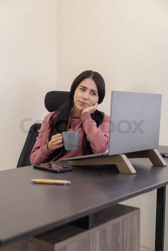 #69318041 woman takes a moment to sip coffee while sitting at her desk, working on a laptop