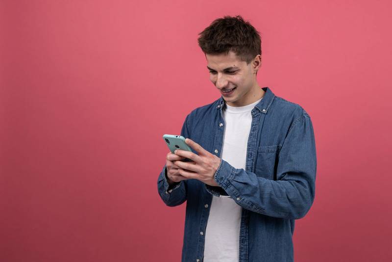 Happy young guy using online smartphone isolated in red background. Smiling and satisfied man texting with phone. 