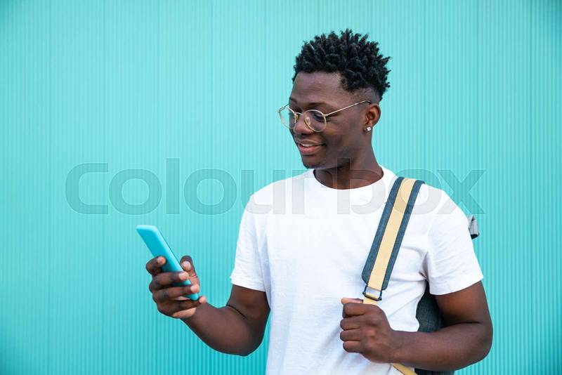 Happy young man smiling and looking his smartphone with a backpack in a turquoise wall - Portrait of a handsome young man