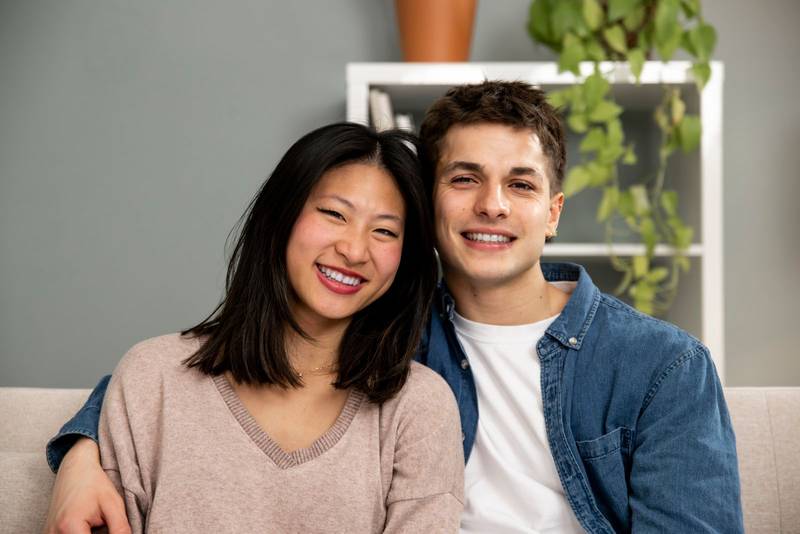 Affectionate smiling young adult couple sitting on sofa at home looking at camera. Carefree man and woman relaxing together in couch at the living room.