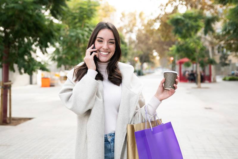 Portrait of beautiful young woman with shopping bags looking at camera with take away coffee and calling with cell telephone 