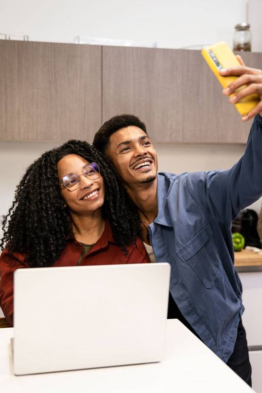 Young adult man and woman taking a photo at their apartment using a computer. Smiling happy diverse couple taking a selfie while working online with laptop in their kitchen at home.