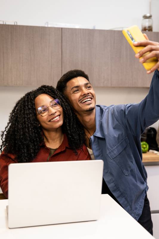 Young adult man and woman taking a photo at their apartment using a computer. Smiling happy diverse couple taking a selfie while working online with laptop in their kitchen at home.