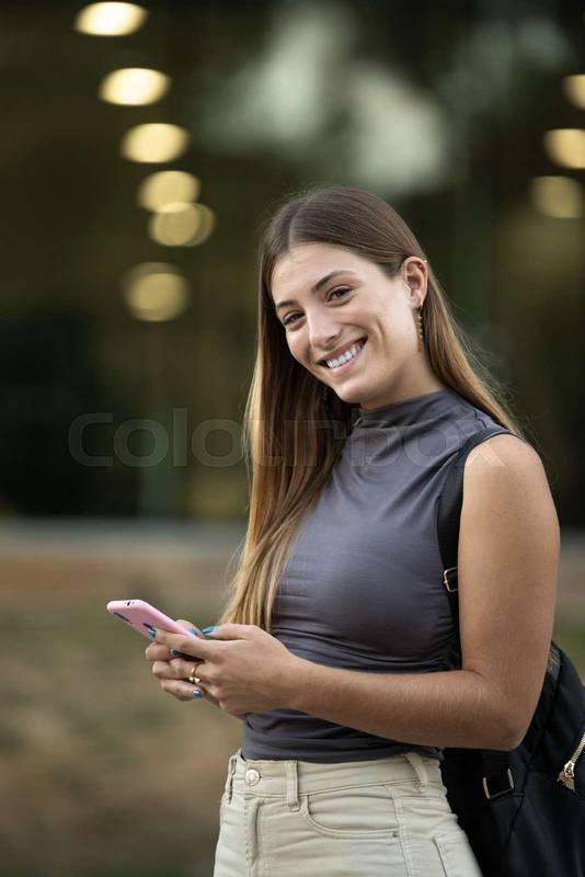 Beautiful cheerful young lady looking at camera texting and standing outside. Joyful blond woman staring at camera in the street while using her phone.