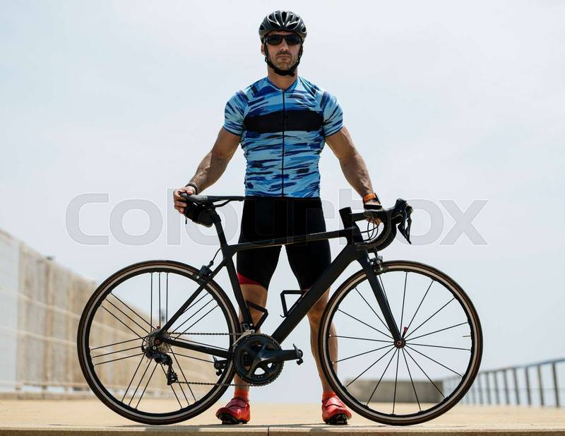Athletic man holding a cycle wearing helmet and sunglasses outside. Cyclist standing with a bike wearing sport clothes in the street.