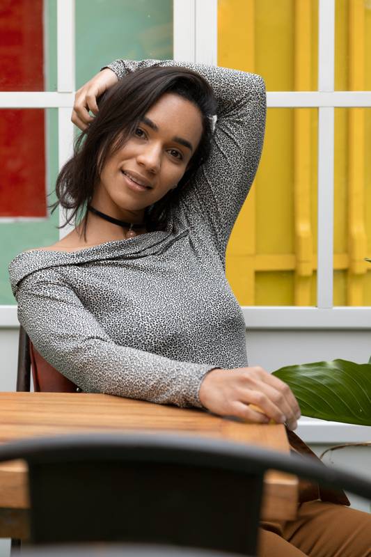 sitting by a wooden table posing with raised arm, wearing casual clothes and a necklace