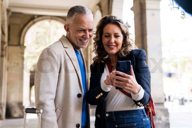 Joyful mid adult couple using smartphone together in the street. Smiling senior husband and wife taking a picture standing in a city.