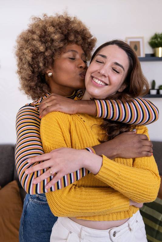 Two lovely and happy homosexual women embracing at home one of them gives a kiss on the cheek to her girlfriend. Beautiful and cheerful multiracial lesbian couple hugging and kissing.