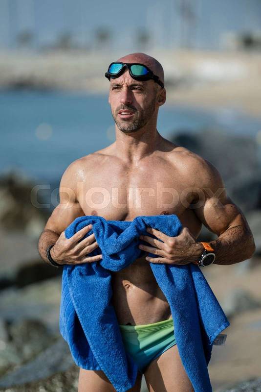 Sexy muscular swimmer toweling off his body with swim glasses at the beach. Fit man with a blue towel after training in the sea wearing speedo.