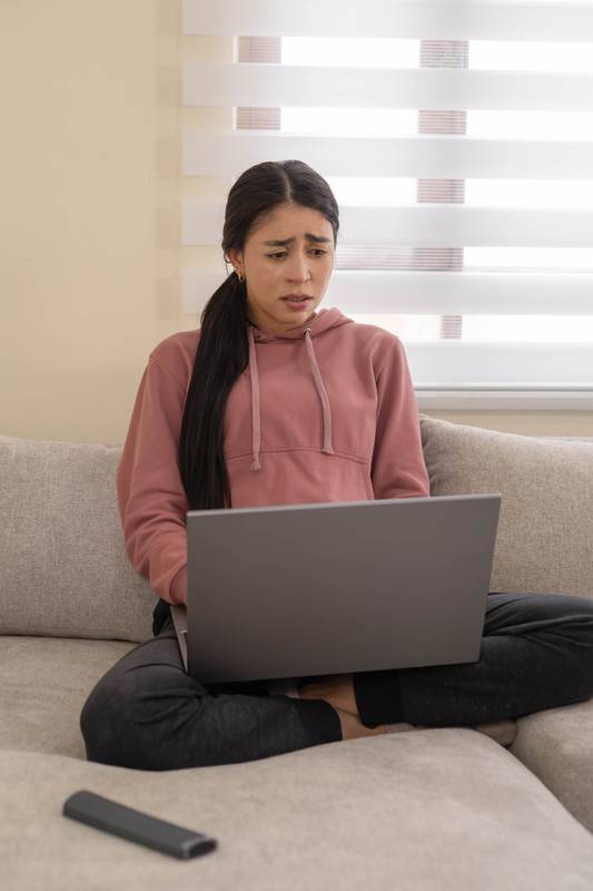 woman sits on a sofa with a concerned expression while using her laptop. Represents stress, bad news, or emotional discomfort