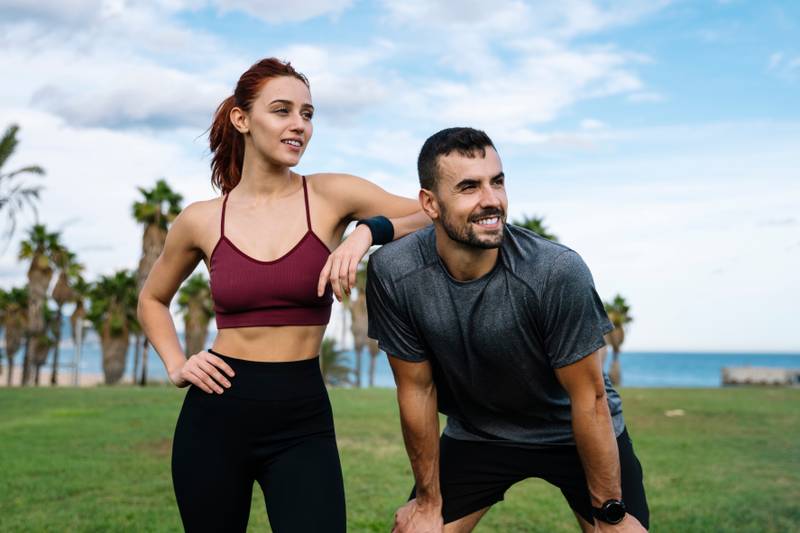 Relaxed young adult couple looking each other after running together in the park. Affectionate satisfied athletic male and female in sportswear training workout outside.