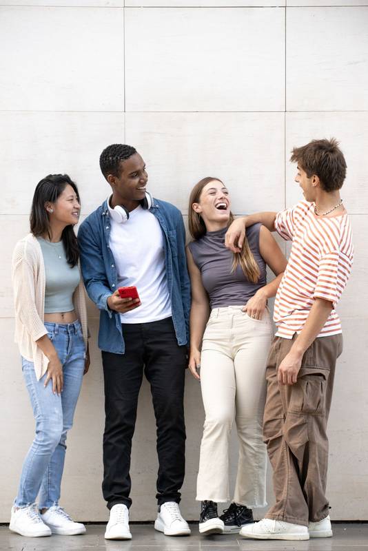 Cheerful men and women having fun outside looking each other Young diverse group of millennials laughing together leaning against an empty wall.