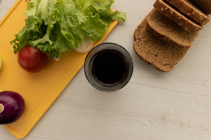 table with a cutting board with fresh vegetables, slices of whole wheat bread and a glass with soda