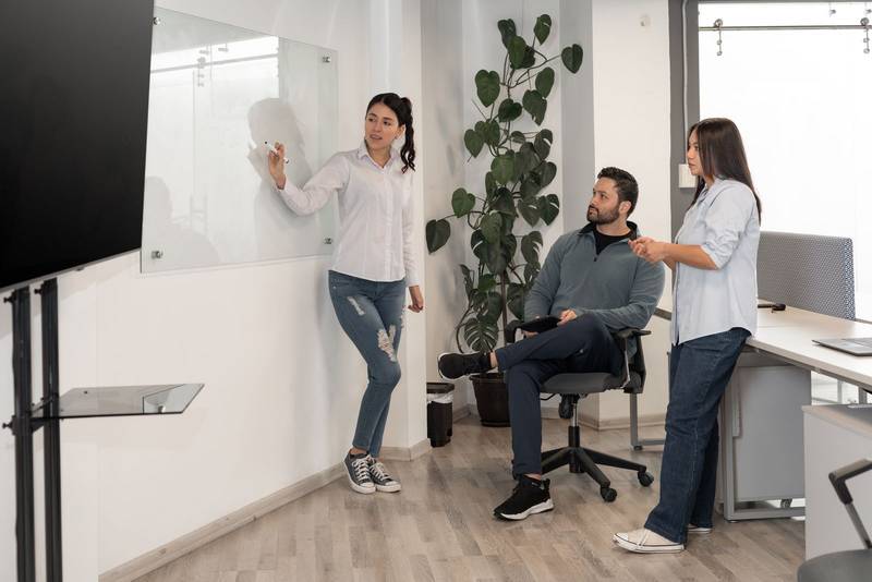 confident woman leads a strategy session, writing on a whiteboard while two team members