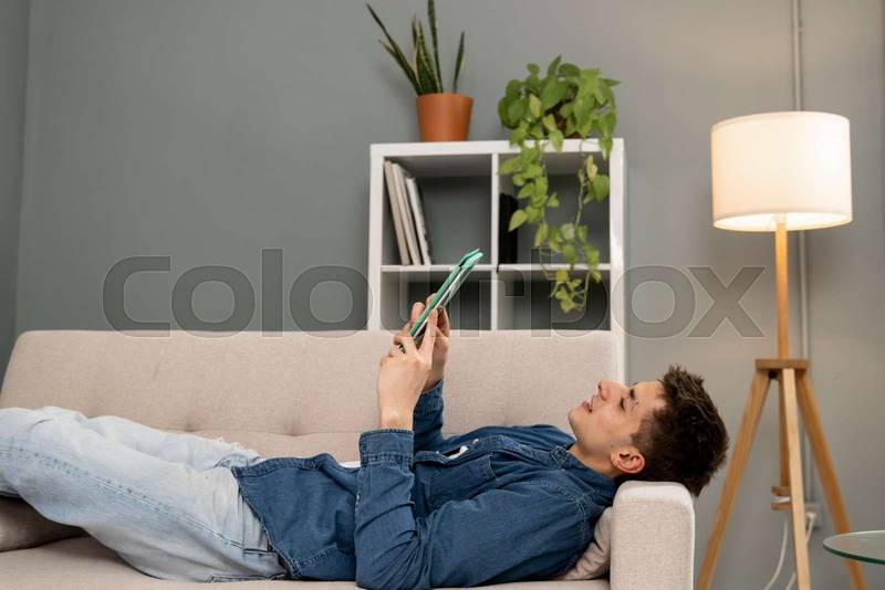 Relaxed young carefree man watching video with a tablet at home. Handsome male resting using with touchpad while sitting on a couch.