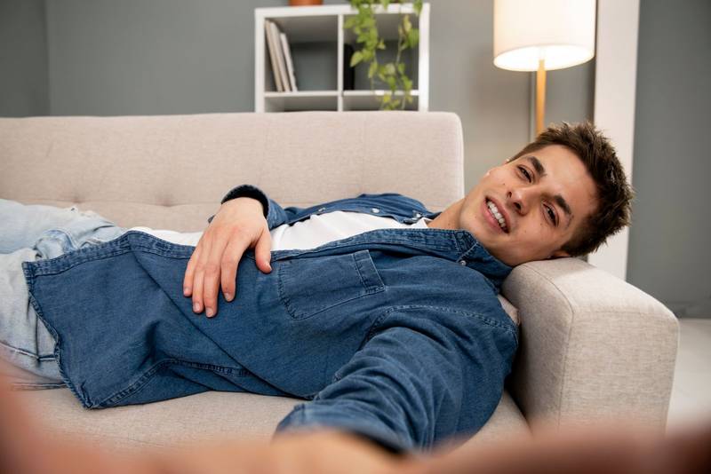 Smiling male laying taking selfie while relaxing on home sofa. Handsome young adult guy resting at home and taking a photo at the living room.