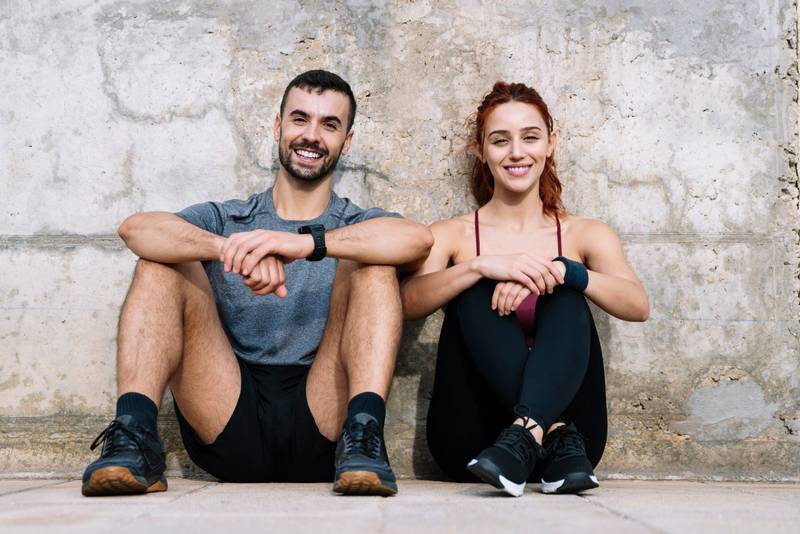 Exhausted young adult couple resting in the floor after running together in the park. Affectionate satisfied athletic male and female sitting in the grass in sportswear training and workout outside. 