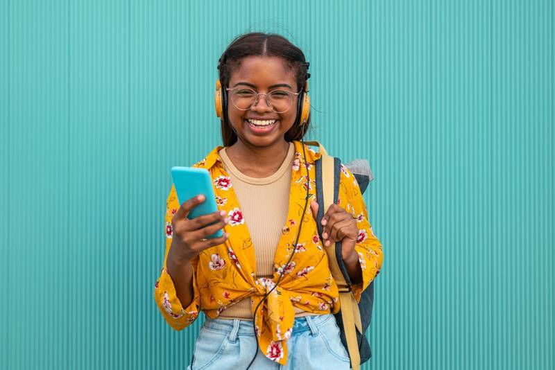 Happy young student smiling and looking at camera with a smartphone, backpack and headphones in a turquoise wall.