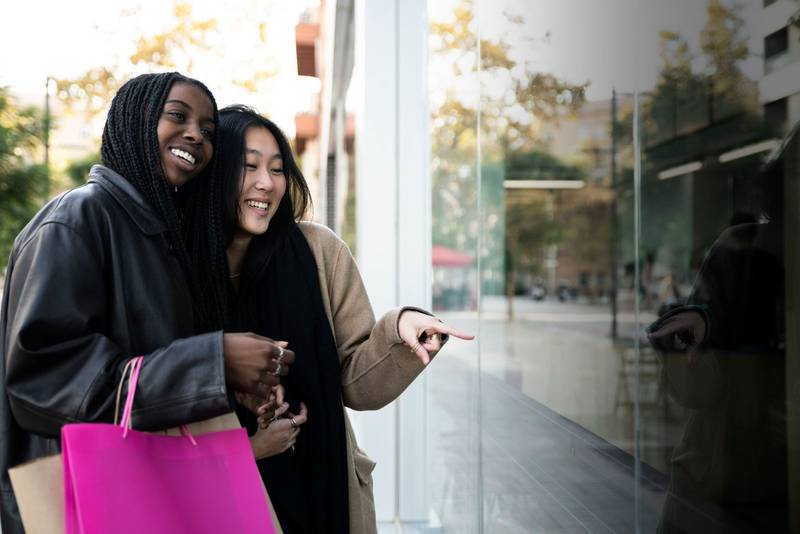 Happy young women holding shopping bags looking to showcase shop - Sale, shopping, consumerism and people concept