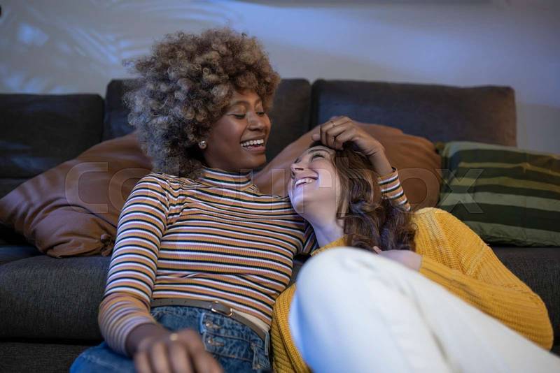 Multiracial lesbian couple hugging and laughing and relaxing in a living room. Smiling gay females couple having fun at home embracing each other.