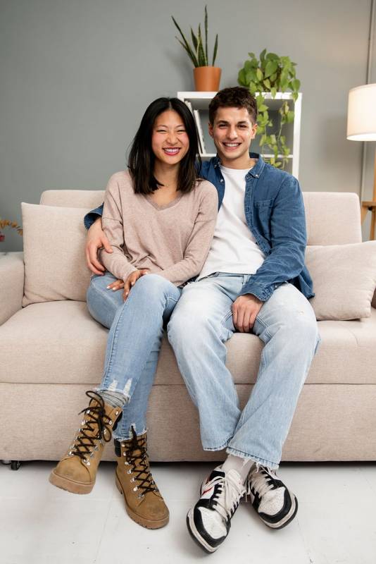 Lovely affectionate smiling young adult couple sitting on sofa at home looking at camera. Diverse carefree man and woman hugging and relaxing together in couch at the living room.