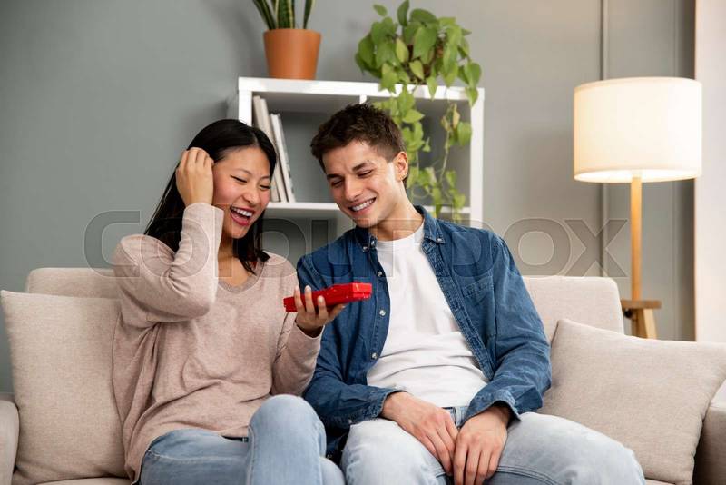 Young diverse couple with mobile phone laughing on sofa. Handsome woman and man sharing smartphone sitting on a couch in their living room.