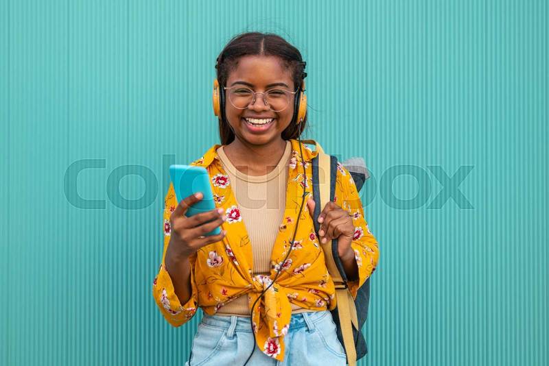 Happy young student smiling and looking at camera with a smartphone, backpack and headphones in a turquoise wall.