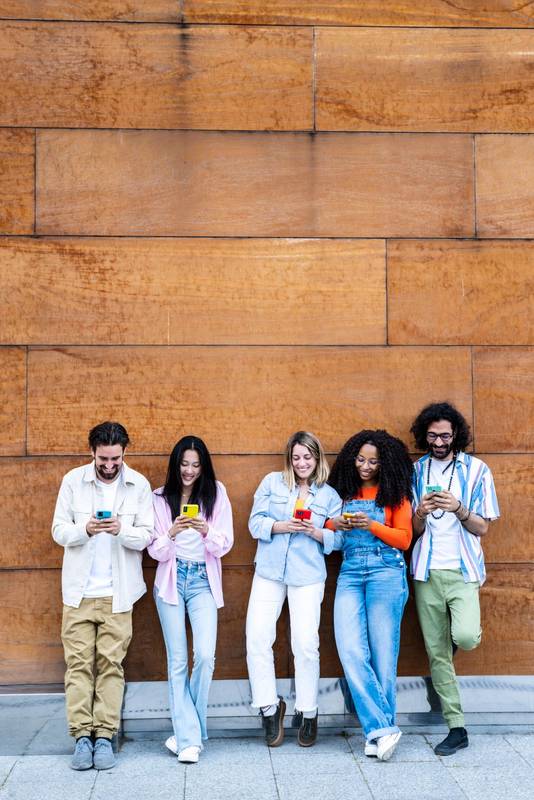 Multiethnic group of friends texting on their smartphones while standing on a brown wall in the street with copy space. Diverse young people smiling and using their phones outside in a city 