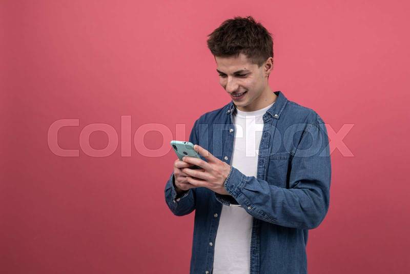 Happy young guy using online smartphone isolated in red background. Smiling and satisfied man texting with phone. 
