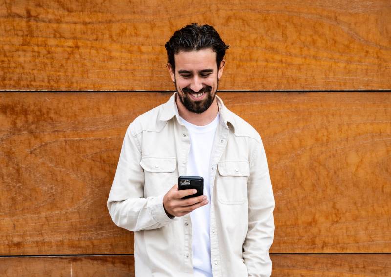 Confident handsome young guy leaning against a wooden wall while texting with his phone. Happy and relaxed man using his smartphone standing on a brown wall outside.