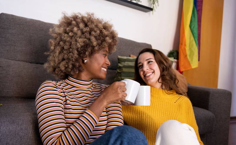 Multiethnic couple taking a coffee break sitting in the floor of a living room. Two cheerful women looking each other while toasting with cups in their apartment.