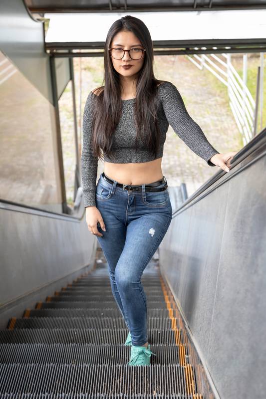 woman with makeup and long hair, wears glasses, jeans with strap and gray top standing on some electric stairs