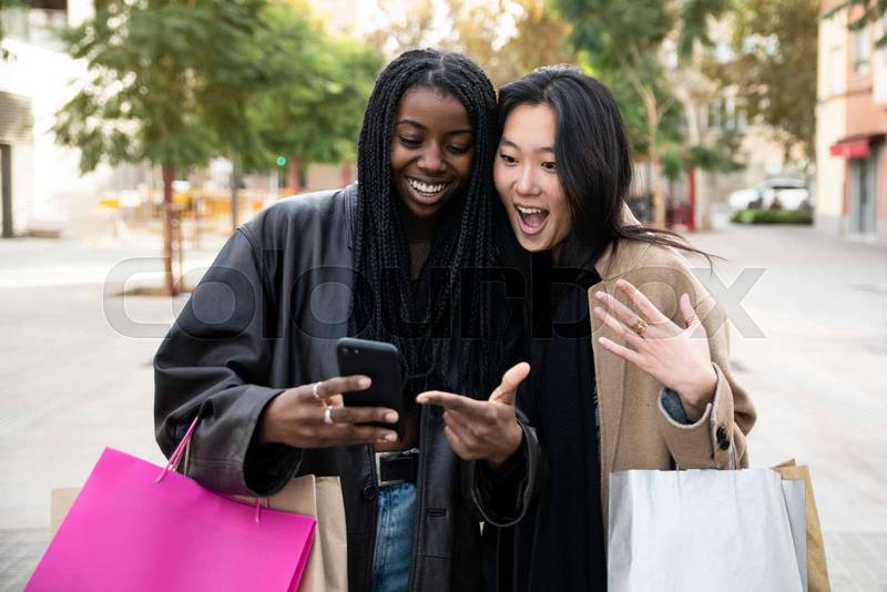 Two surprised young beautiful women holding shopping bags and looking smartphone. Cheerful friends amazed with her mobile phone. 