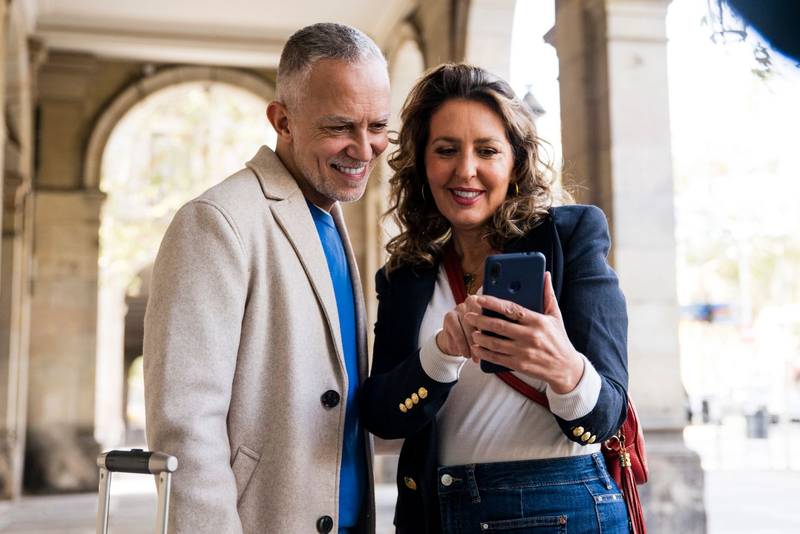Joyful mid adult couple using smartphone together in the street. Smiling senior husband and wife taking a picture standing in a city.