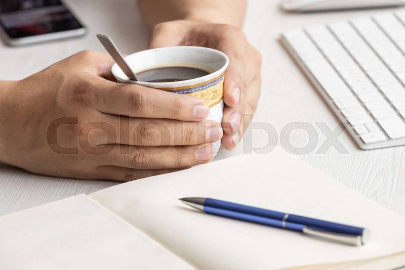 workplace with a person's hands holding a cup with hot coffee, next to it an open notebook