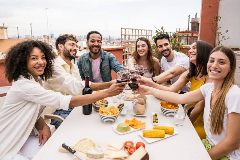 Happy group of friends toasting with wine glasses in a terrace looking at camera. Diverse group of young people drinking in a rooftop dinner staring at camera.