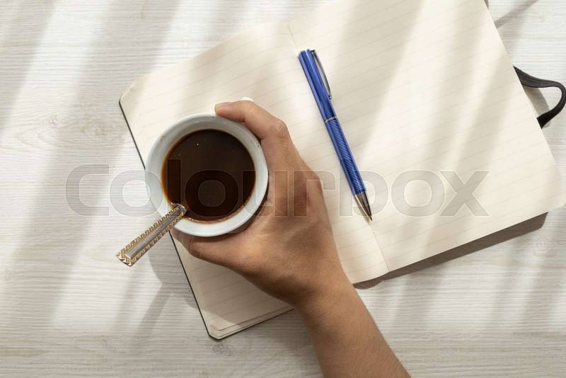 notebook with open blank sheets, on top of it a hand holding a cup with coffee next to a pen