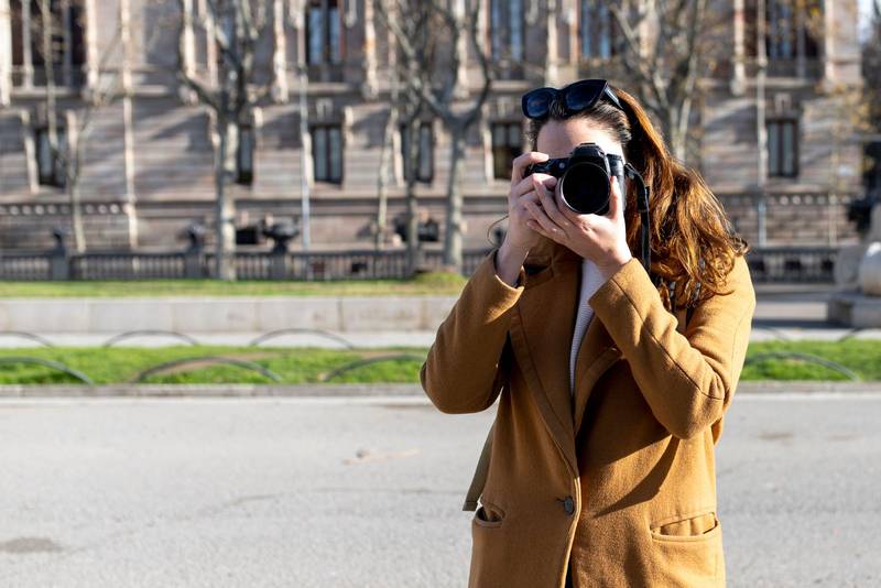 Young female tourist taking photos in street with camera. Woman photographer covering her face with a camera
