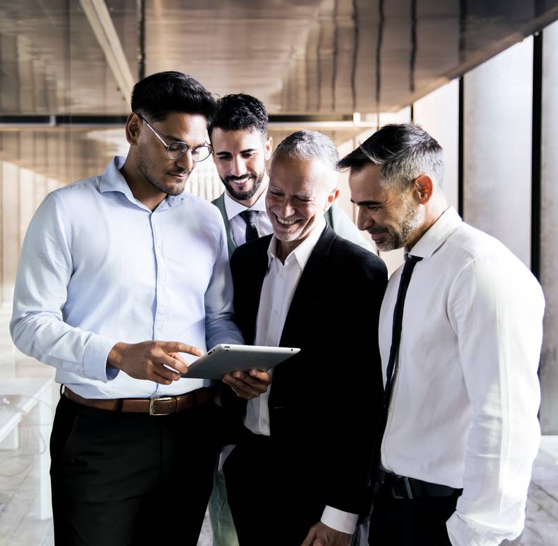 Diverse group of businessmen checking a tablet in an office corridor. Young executive holding a device in a meeting with senior colleagues standing at office.
