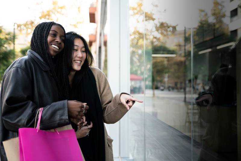 Happy young women holding shopping bags looking to showcase shop - Sale, shopping, consumerism and people concept