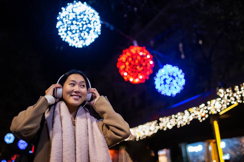 Young happy woman wearing headphones with christmas lights behind. Joyful girl listening to music at the street during winter.