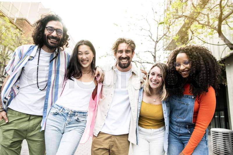 Diverse group of cheerful millennial friends standing together hugging each other in street. Cheerful multiethnic young carefree friends laughing and having fun looking at camera.