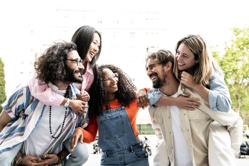 Multiracial group of friends giving piggyback ride and looking each other. Diverse young women and men hugging each other laughing and carefree staring at each other outside.