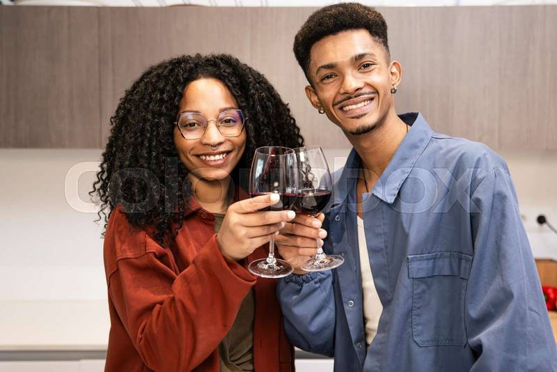 Happy young man and woman drinking with glasses in home. Cheering diverse couple toasting with wine in kitchen.
