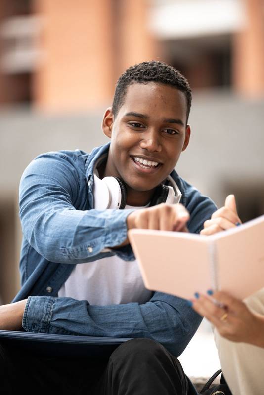 Young handsome guy wearing headphones pointing a notebook.Confident student sitting and reading notes with a colleague.