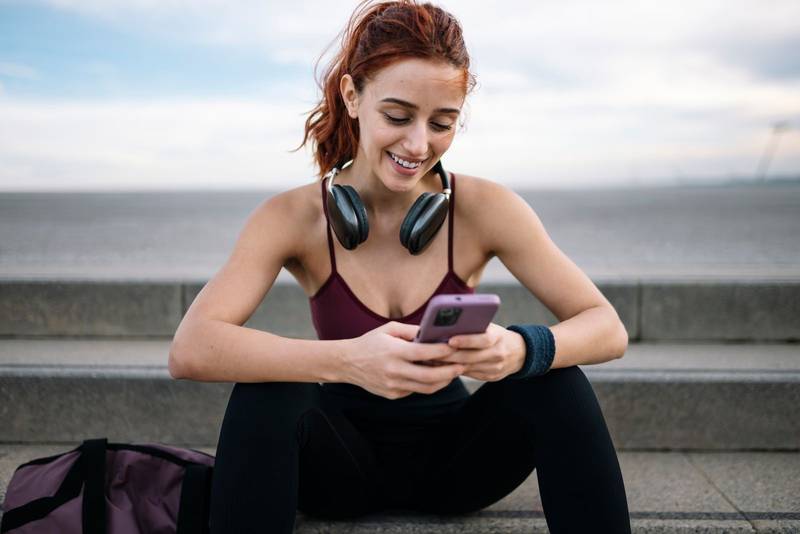 Joyful sports woman typing with phone wearing headphones sitting outside. Young adult fit athletic female in sportswear texting with smartphone