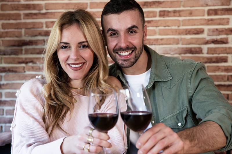  Cheerful male and female drinking with glasses at home. Joyful young adult couple toasting with wine and looking at camera.